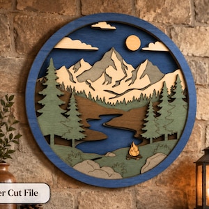 Peut inclure: Une décoration murale circulaire en bois multicouche représentant un paysage de montagne avec une rivière bleue, des pins verts, un soleil jaune et un petit feu de camp. Le design inclut des nuages et un cadre circulaire bleu foncé.