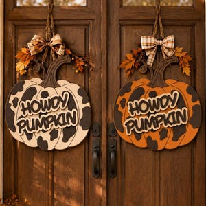 May include: Two wooden pumpkin-shaped door signs hanging on a double door. One sign is white with black cow print spots, and the other is orange with black cow print spots. Both feature the text HOWDY PUMPKIN, a dark brown stem, and a plaid bow.
