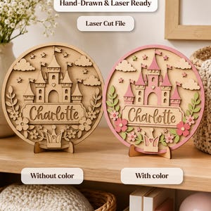 Op de afbeelding: Twee ronde houten borden met een lasergesneden kasteel, wolken, sterren, bloemen en een kroon, met de naam Charlotte in het midden gegraveerd. Het ene bord is van natuurlijk hout, het andere is beschilderd in roze en groen. Tekst: Hand-Drawn & Laser Ready, Laser Cut File, Without color, With color.