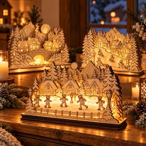 Op de afbeelding: Drie verlichte houten diorama's met winterse dorpsscènes. Elk stuk toont gedetailleerde, lasergesneden silhouetten van huizen, dennenbomen en figuren die schaatsen of sleeën, allemaal verlicht door een warme gloed van binnenuit.