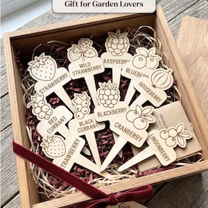 May include: A wooden box containing light-colored wooden plant markers with engraved fruit illustrations and names including Strawberry, Wild Strawberry, Raspberry, Blueberry, Gooseberry, Red Currant, Black Currant, Blackberry, Cranberry, and Lingonberry.