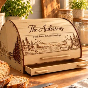 Peut inclure: Une boîte à pain en bois clair avec un couvercle coulissant orné d'une gravure laser représentant un paysage de montagne et de lac. Le texte indique The Andersons et Fresh Bread & Cozy Mornings. Elle possède un tiroir à miettes.