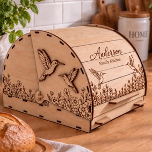 Könnte beinhalten: Ein hellfarbiger Brotkasten aus Holz mit gewölbtem Deckel, verziert mit lasergravierten Kolibris und Blumenmustern. Der Text "Anderson" und "Family Kitchen" ist auf der Vorderseite eingraviert. Das Gehäuse hat eine Schiebetür.