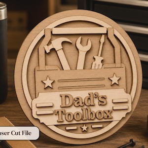 May include: A wooden, circular sign with a toolbox design. The sign features a hammer, wrench, and screwdriver, with the words "Dad's Toolbox" and star accents. A black tumbler with "DAD FUEL" is visible.