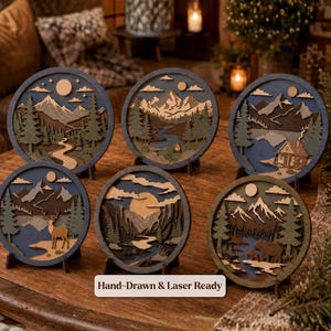 Peut inclure: Six décorations murales circulaires en bois superposé représentant des paysages de montagne avec des arbres, des rivières et des animaux comme un cerf et un ours. Chaque pièce est sur un support. Le texte Hand-Drawn & Laser Ready est inscrit.