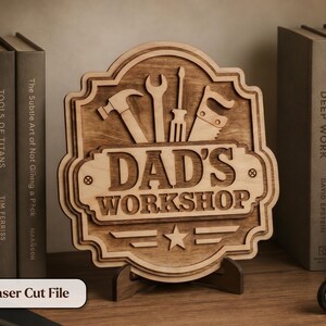 May include: Wooden sign with the words "DAD'S WORKSHOP" and tools like a hammer, wrench, screwdriver, and saw. The sign is laser cut and stands on a wooden base. The sign is displayed on a wooden surface.
