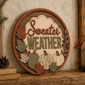 Pode incluir: Uma placa de parede circular de madeira com o texto "Sweater WEATHER" em letras sobrepostas. O design inclui três abóboras, uma com estampa de leopardo, rodeadas por folhas de outono e bagas em tons suaves.