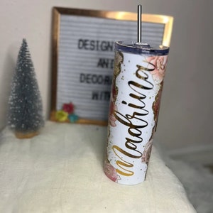 K&ouml;nnte beinhalten: Ein wei&szlig;er Becher mit Blumenmuster und goldenem Glitzer. Auf dem Becher steht das Wort "Magnolia" in goldener Kursivschrift.
