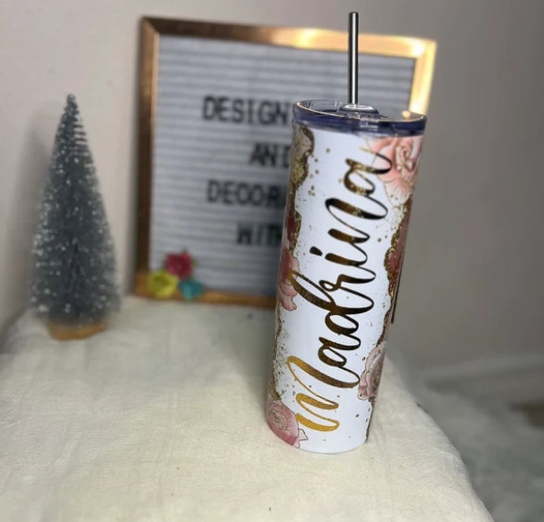 K&ouml;nnte beinhalten: Ein wei&szlig;er Becher mit Blumenmuster und goldenem Glitzer. Auf dem Becher steht das Wort "Magnolia" in goldener Kursivschrift.