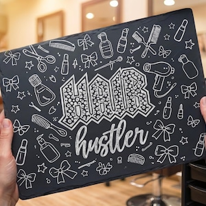 Könnte beinhalten: Eine schwarze Tafel mit weißen Illustrationen von Friseurwerkzeugen und den Worten "HAIR hustler". Das Schild wird von zwei Händen gehalten, mit einem Saloninterieur im Hintergrund.
