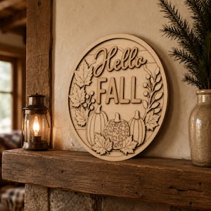 May include: A circular wooden wall sign featuring the text Hello Fall in a script and block font. The design includes layered cutouts of three pumpkins, one with a leopard print pattern, surrounded by maple leaves and sprigs on a plank background.