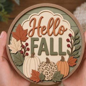 May include: A circular wooden sign featuring the text Hello FALL in layered script and block lettering. The design includes cutouts of three pumpkins, one with a leopard print pattern, surrounded by autumn leaves and red berries in shades of green, orange, and cream.