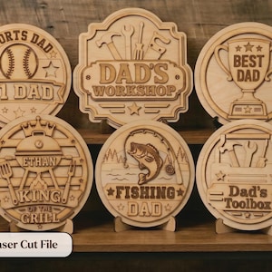 May include: A collection of laser-cut wooden signs with various themes for Father's Day. Designs include "Sports Dad," "Dad's Workshop," "Best Dad," "King of the Grill," "Fishing Dad," and "Dad's Toolbox." Each sign is displayed on a wooden stand.