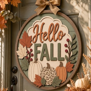 May include: A circular wooden door sign featuring the text Hello FALL in layered script and block lettering. The design includes three pumpkins, one with a leopard print pattern, surrounded by autumn leaves and berries in shades of orange, green, and cream, topped with a burlap bow.