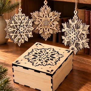 May include: Three laser-cut wooden snowflake ornaments with intricate layered designs hang from twine strings above a square wooden box featuring a matching black snowflake cutout on its lid and black notched corner detailing.