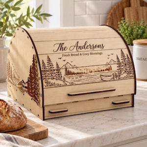 Puede incluir: Una panera de madera clara con tapa enrollable que presenta un paisaje de montaña y lago grabado con láser. El texto "The Andersons" y "Fresh Bread & Cozy Mornings" está grabado en la parte frontal. Incluye un cajón extraíble en la base.