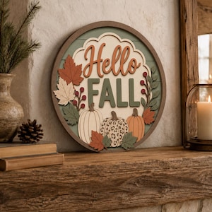 May include: A circular wooden wall sign featuring the text Hello Fall in raised lettering. The design includes three pumpkins, one with a leopard print pattern, surrounded by layered autumn leaves and berries in shades of orange, green, and cream.
