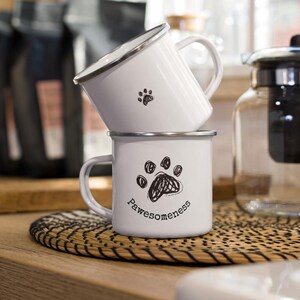 Pode incluir: Duas canecas de esmalte branco empilhadas sobre um tapete tecido. A caneca superior tem uma pequena pegada de pata, enquanto a caneca inferior apresenta uma pegada de pata maior com a palavra "Pawesomeess" abaixo. Ambas as canecas têm bordas e alças prateadas.