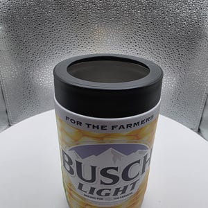 Puede incluir: Un aislante de bebidas cil&iacute;ndrico con un borde negro y un dise&ntilde;o impreso en blanco y amarillo. El dise&ntilde;o presenta las palabras "BUSCH LIGHT" y "FOR THE FARMERS". El aislante est&aacute; dise&ntilde;ado para mantener las bebidas fr&iacute;as.