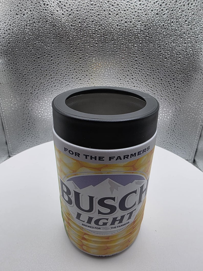 Puede incluir: Un aislante de bebidas cil&iacute;ndrico con un borde negro y un dise&ntilde;o impreso en blanco y amarillo. El dise&ntilde;o presenta las palabras "BUSCH LIGHT" y "FOR THE FARMERS". El aislante est&aacute; dise&ntilde;ado para mantener las bebidas fr&iacute;as.