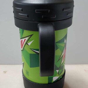 May include: Green and black insulated travel mug with a black handle and a clear lid with a spout. The mug has a Mountain Dew logo and a geometric pattern.