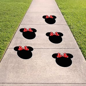 May include: Five black Minnie Mouse head-shaped decals with red and white polka dot bows are arranged on a concrete sidewalk. The decals are spaced apart, creating a playful and whimsical design on the walkway.