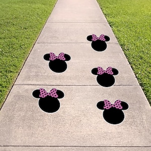 May include: Five black Minnie Mouse head-shaped sidewalk decals with pink polka dot bows are arranged on a concrete walkway. The decals are spaced apart, leading the eye down the path.