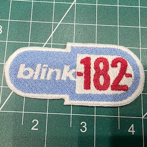 May include: Embroidered patch featuring the text "blink-182" in white and red. The patch has a light blue background with a white border. The patch is in the shape of a pill.