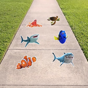 May include: Colorful character cutouts from the animated film "Finding Nemo" and "Finding Dory" are arranged on a concrete walkway. The characters include a shark, clownfish, octopus, sea turtle, and blue tang.