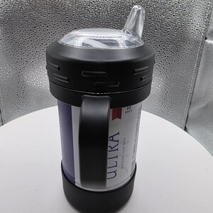 Pode incluir: Um copo t&eacute;rmico preto com uma al&ccedil;a, segurando uma lata de cerveja Ultra. O copo tem uma tampa transparente e angular para beber. A lata &eacute; vis&iacute;vel atrav&eacute;s da abertura do copo, com o logotipo e o texto Ultra em branco.