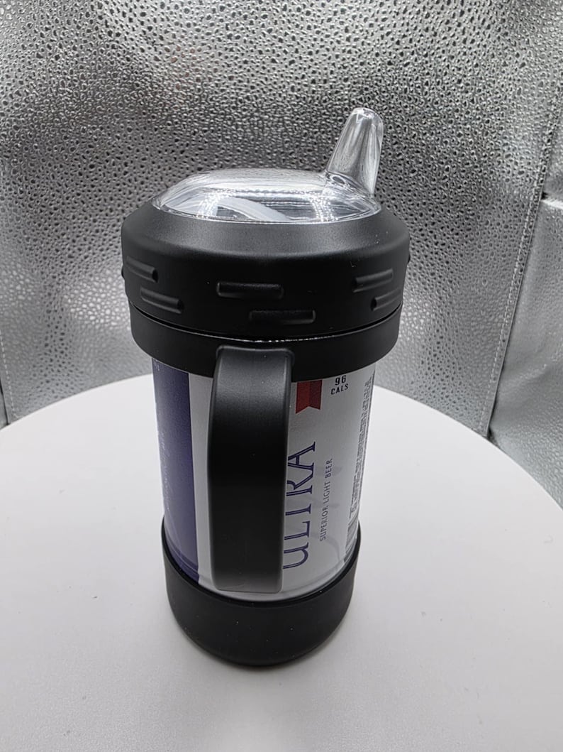 Pode incluir: Um copo t&eacute;rmico preto com uma al&ccedil;a, segurando uma lata de cerveja Ultra. O copo tem uma tampa transparente e angular para beber. A lata &eacute; vis&iacute;vel atrav&eacute;s da abertura do copo, com o logotipo e o texto Ultra em branco.