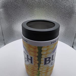 Puede incluir: Un aislante de bebidas cil&iacute;ndrico con tapa negra y banda blanca. El cuerpo presenta un dise&ntilde;o de mazorca de ma&iacute;z con las palabras "Ranch Buds" impresas. El fondo es blanco neutro.