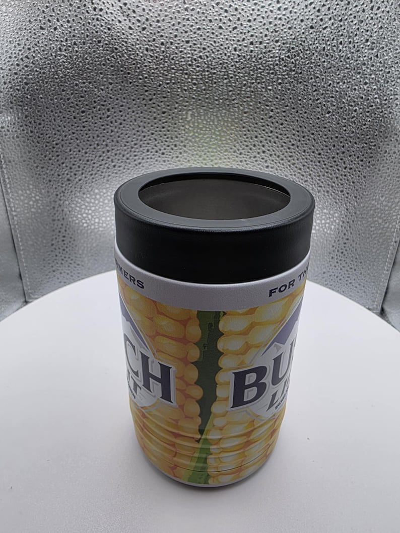 Puede incluir: Un aislante de bebidas cil&iacute;ndrico con tapa negra y banda blanca. El cuerpo presenta un dise&ntilde;o de mazorca de ma&iacute;z con las palabras "Ranch Buds" impresas. El fondo es blanco neutro.