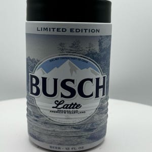May include: A cylindrical can with a black lid and a blue and white design. The can features the text "LIMITED EDITION" and "BUSCH Latte" with the words "BREWED FOR AMERICA'S HEARTLAND". The can contains 12 fl oz of beer.