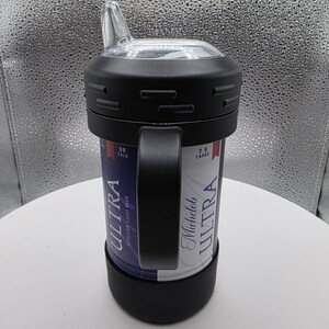 Pode incluir: Um copo t&eacute;rmico preto com uma al&ccedil;a, projetado para conter uma lata de cerveja Michelob Ultra. Possui uma tampa de pl&aacute;stico transparente com bico. A lata &eacute; vis&iacute;vel, com as palavras "ULTRA" e "SUPERIOR LIGHT BEER" impressas.