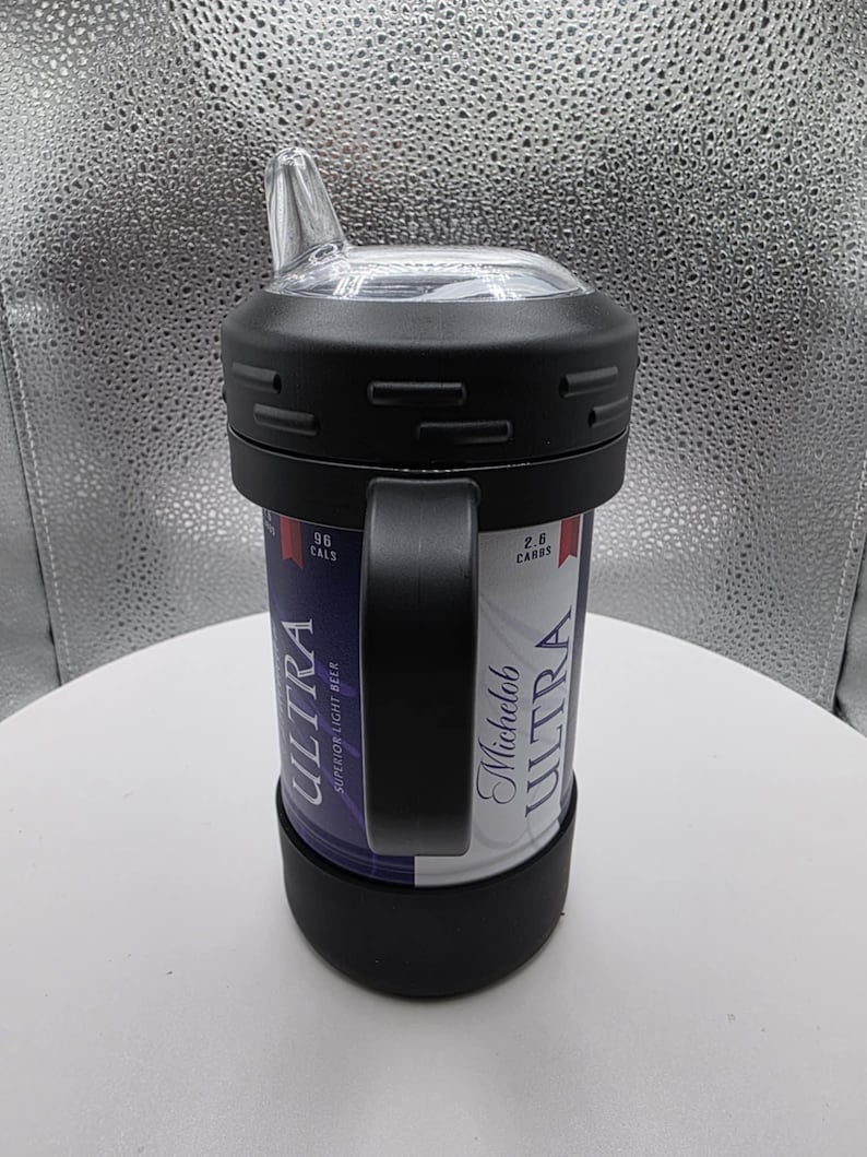 Pode incluir: Um copo t&eacute;rmico preto com uma al&ccedil;a, projetado para conter uma lata de cerveja Michelob Ultra. Possui uma tampa de pl&aacute;stico transparente com bico. A lata &eacute; vis&iacute;vel, com as palavras "ULTRA" e "SUPERIOR LIGHT BEER" impressas.