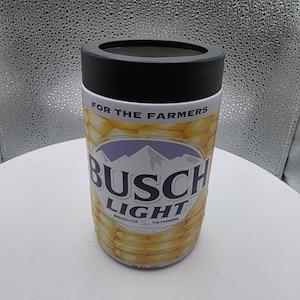 Puede incluir: Un aislante de bebidas cil&iacute;ndrico con una tapa negra y un dise&ntilde;o con tem&aacute;tica de ma&iacute;z. Las palabras "BUSCH LIGHT" se muestran de forma destacada, con "BREWED FOR THE FARMERS" impreso debajo. El aislante est&aacute; dise&ntilde;ado para contener una lata de 355 ml.