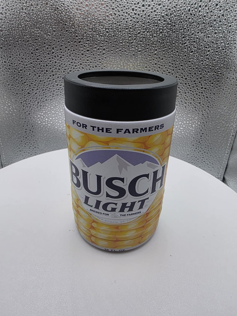 Puede incluir: Un aislante de bebidas cil&iacute;ndrico con una tapa negra y un dise&ntilde;o con tem&aacute;tica de ma&iacute;z. Las palabras "BUSCH LIGHT" se muestran de forma destacada, con "BREWED FOR THE FARMERS" impreso debajo. El aislante est&aacute; dise&ntilde;ado para contener una lata de 355 ml.