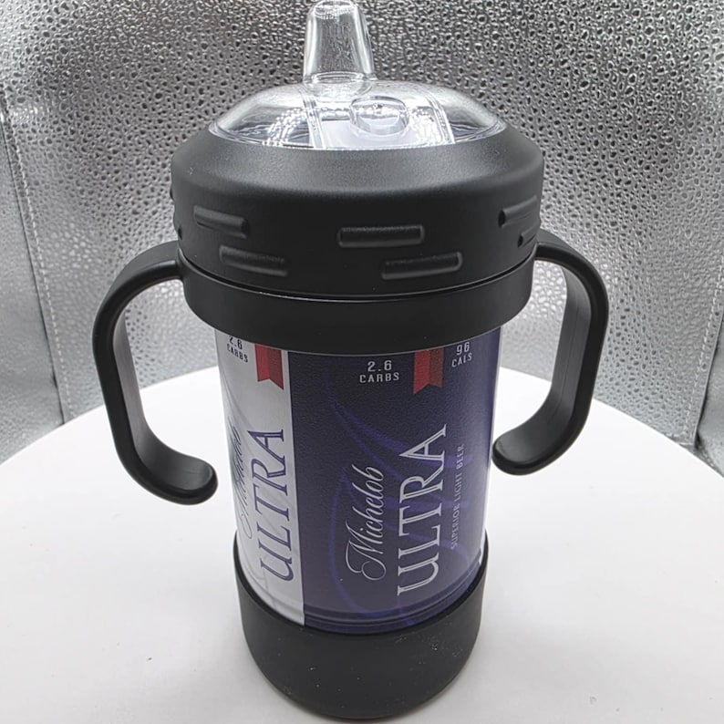 Pode incluir: Uma caneca preta com duas al&ccedil;as, uma parte superior transparente e uma base preta. A caneca &eacute; embrulhada com um r&oacute;tulo de lata de cerveja Michelob Ultra. O r&oacute;tulo &eacute; azul e branco com as palavras "ULTRA" e "Michelob Ultra".