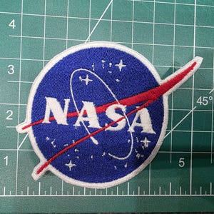 May include: Embroidered patch featuring the NASA logo. The patch has a blue background with white text and a red swoosh. The NASA logo includes a stylized orbit and stars.