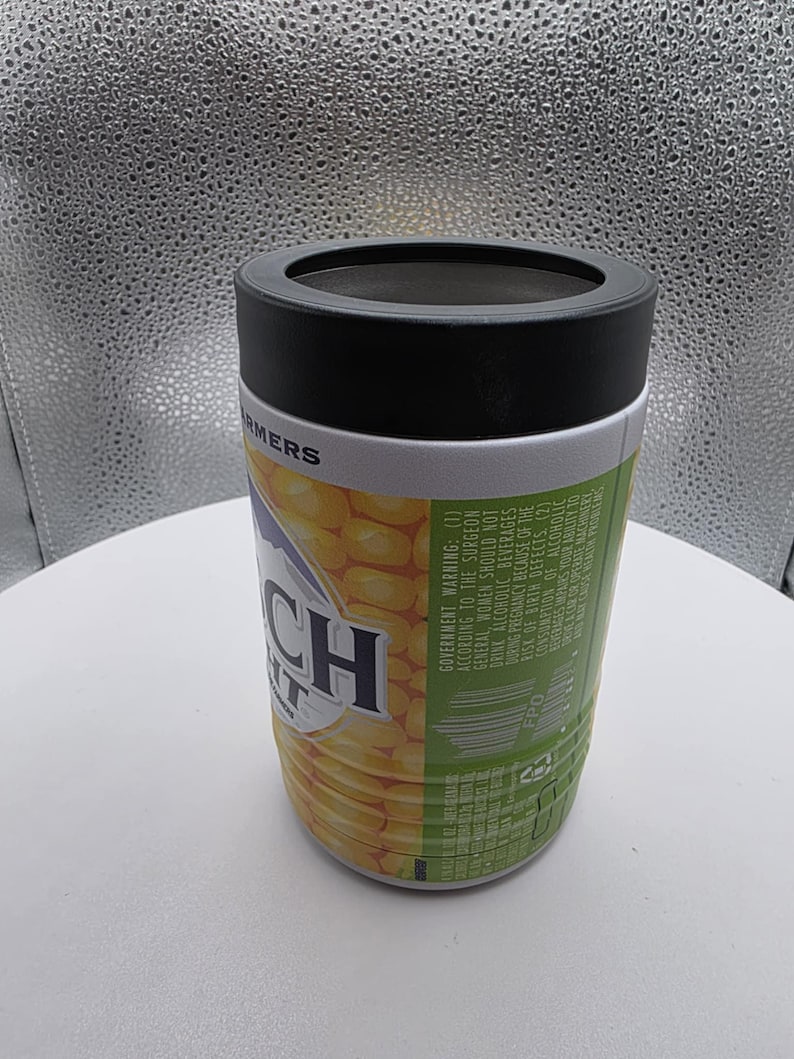 Puede incluir: Un enfriador de latas cil&iacute;ndrico con una tapa negra y un dise&ntilde;o con mazorcas de ma&iacute;z. El texto "Ranch" es visible en la parte delantera. El enfriador tiene una banda blanca con la palabra "FARMERS" impresa.