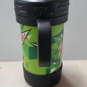 May include: Green and black travel mug with a black handle and a clear lid with a spout. The mug has a Mountain Dew logo and a geometric pattern.