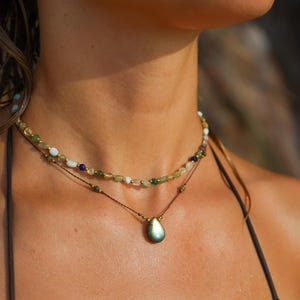 Könnte beinhalten: Eine mehrreihige Halskette mit einem tropfenförmigen Anhänger und Perlensträngen in Grün, Weiß und Blau. Die Halskette wird mit einem passenden Ohrring getragen. Der Schmuck wird auf der Brust einer Person präsentiert.