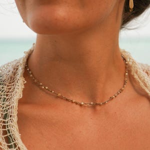 Può includere: Una delicata collana con piccole perline di colore chiaro e un paio di orecchini a goccia abbinati. I gioielli sono indossati su uno sfondo di un capo di abbigliamento chiaro a trama aperta e una vista sfocata dell'oceano.