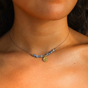 Könnte beinhalten: Zarte Halskette mit kleinen, hellblauen Perlen und goldenen Akzenten. Ein goldener Anhänger mit Sonnenmuster hängt unter den Perlen. Die Halskette ist an einem dünnen, neutralfarbenen Band befestigt.