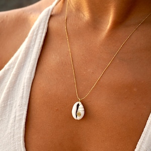 Op de afbeelding: Een goudkleurige ketting met een witte schelpenhanger. De hanger heeft een spiraalvormig ontwerp en goudkleurige metalen accenten. De ketting wordt op de borst van een persoon getoond.