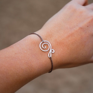 Könnte beinhalten: Silberfarbenes Drahtarmband mit Spiraldesign. Das Armband ist an einem dunkelbraunen Kordelband befestigt. Der minimalistische Schmuck ist ein stilvolles Accessoire.