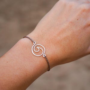 Op de afbeelding: Zilverkleurige spiraalvormige bedelarmband. De bedel is verbonden met een dun, bruin koord. De armband wordt om een pols gedragen. De achtergrond is wazig.