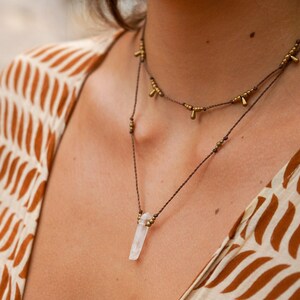 May include: A delicate necklace with a clear quartz crystal pendant and gold-toned beads. The necklace is made of brown cord and has a simple, minimalist design.