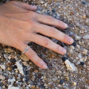 Könnte beinhalten: Ein silberner Ring mit kleinen, runden, grauen Steinen, die in einem filigranen Kettenmuster eingefasst sind. Der Ring wird an einem Finger getragen, der auf einem Bett aus kleinen, grauen, weißen und braunen Kieselsteinen liegt.